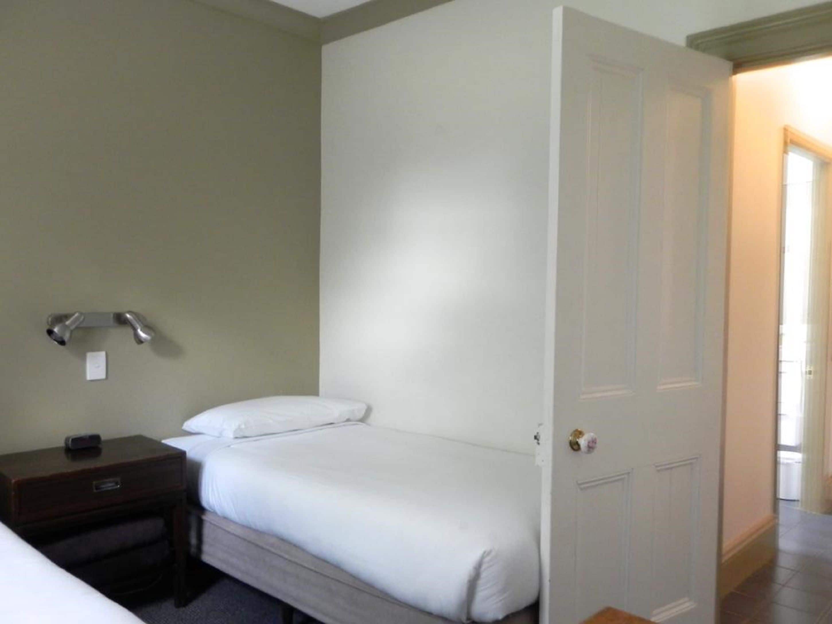 A hotel room with a single bed, a bedside table, a nightstand, and a door leading to another room.