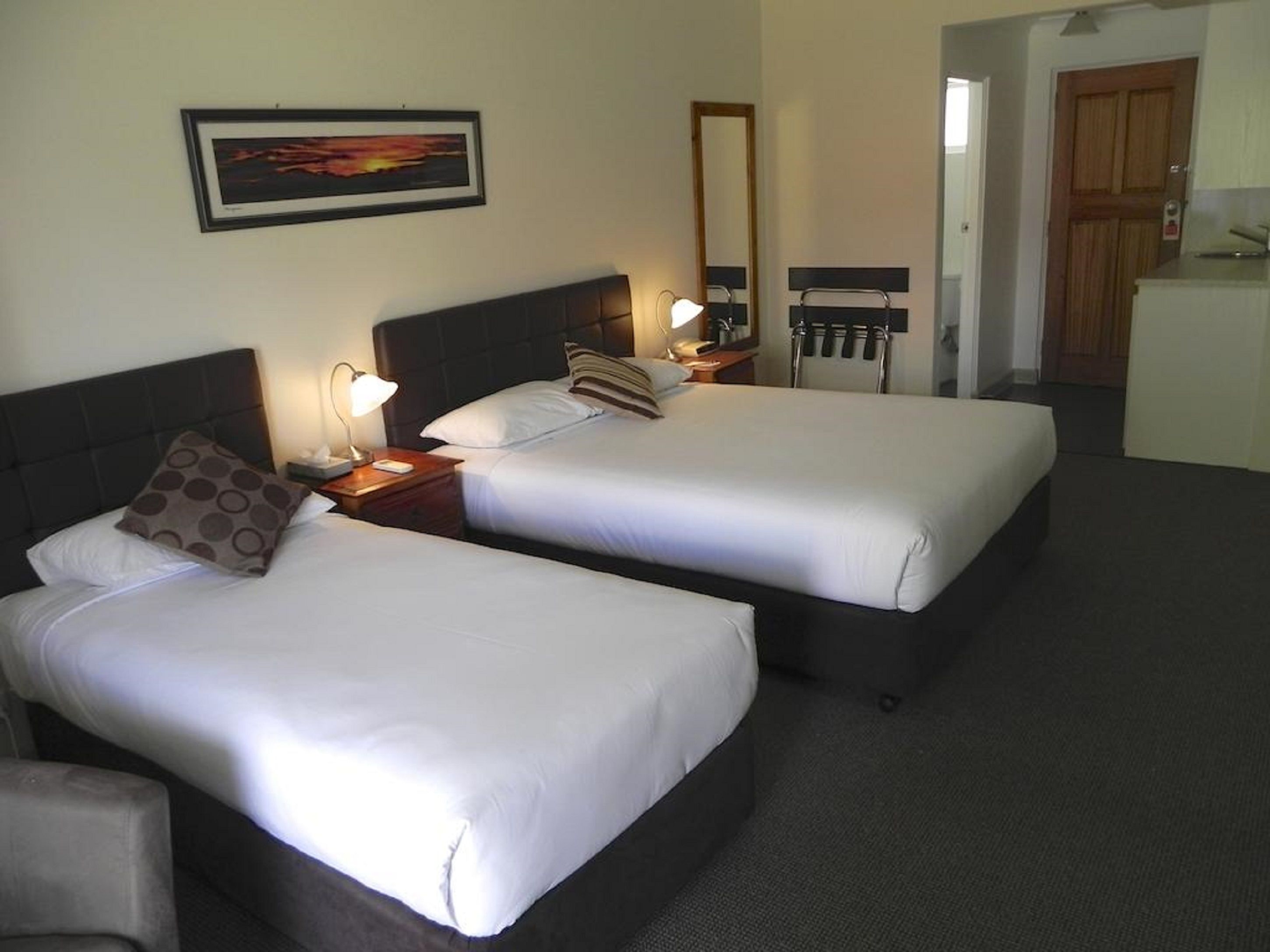 A hotel room with two beds, a nightstand with a lamp, a mirror, and a view of a kitchen area.