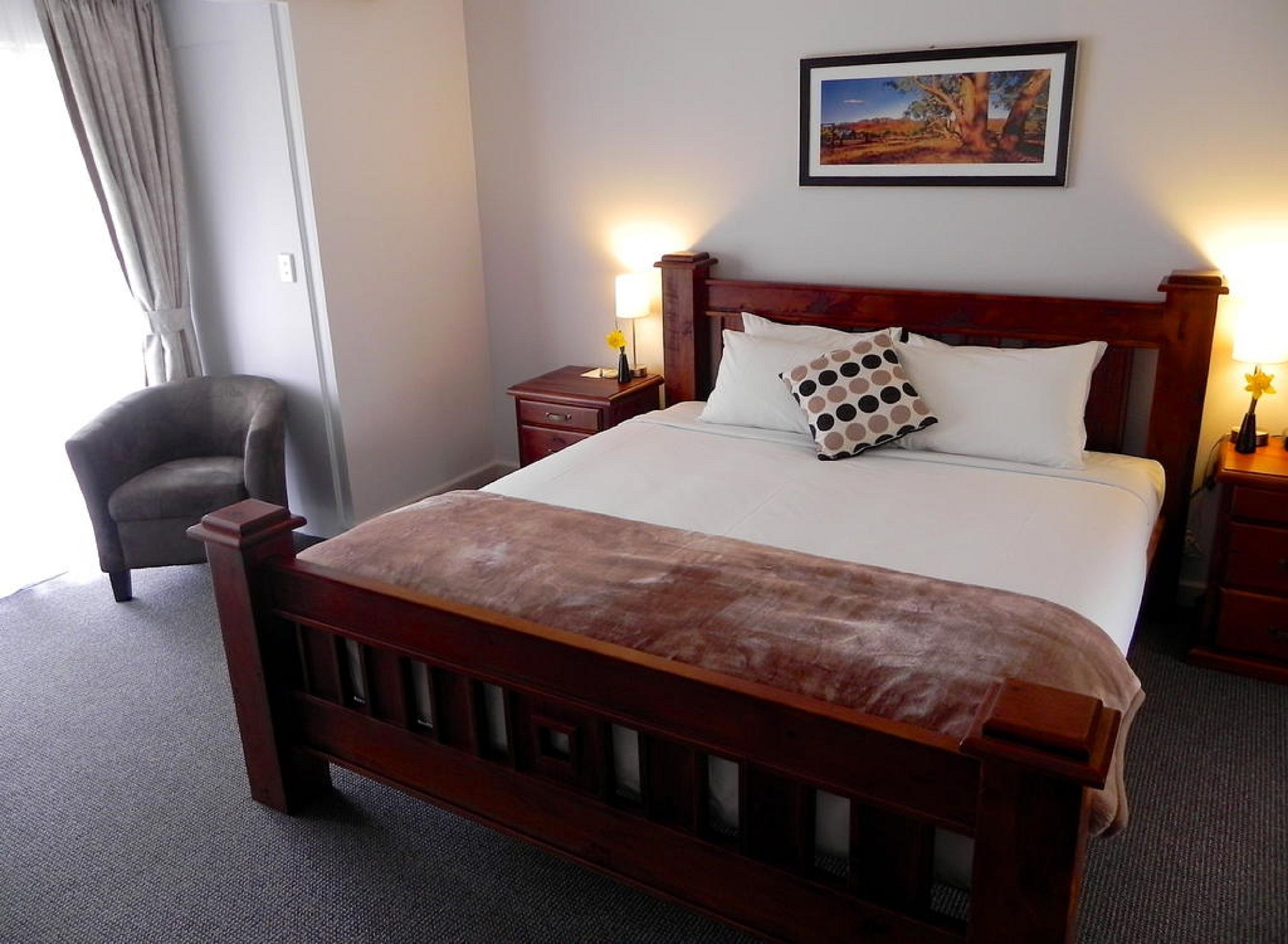 Een hotelkamer met een houten bed, een nachtkastje met een lamp, een stoel en een schilderij aan de muur.