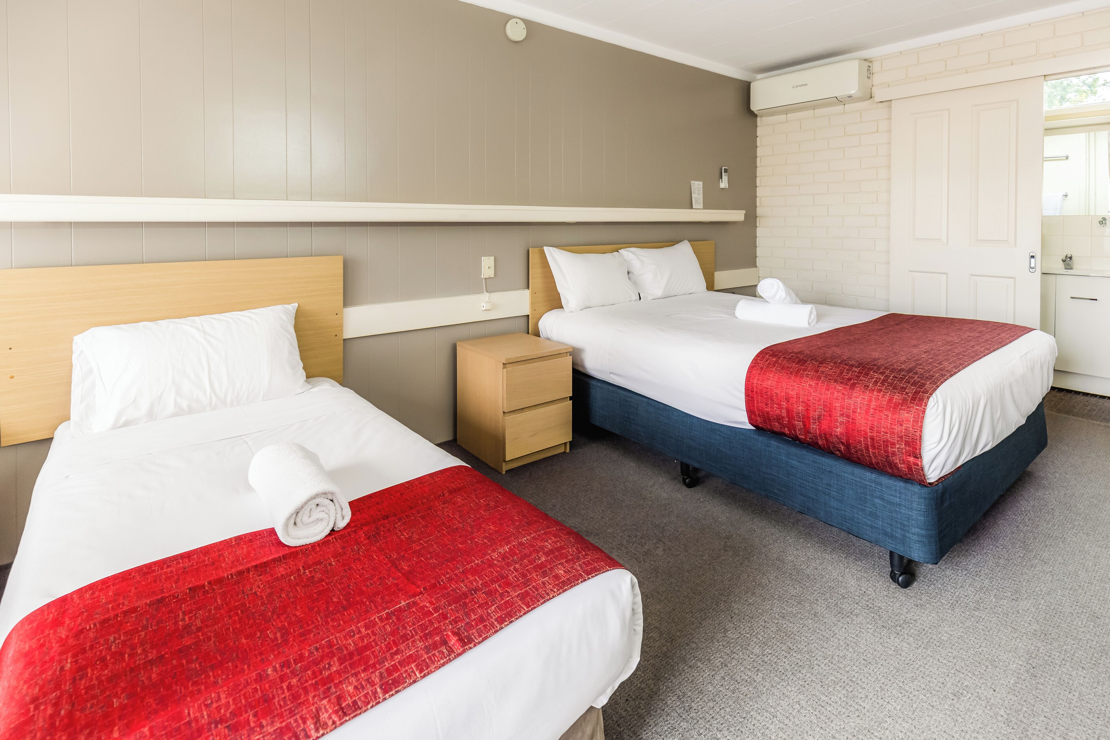 Two single beds with wooden headboards, a small wooden nightstand, a white wall with a shelf, and a door with a mirror.