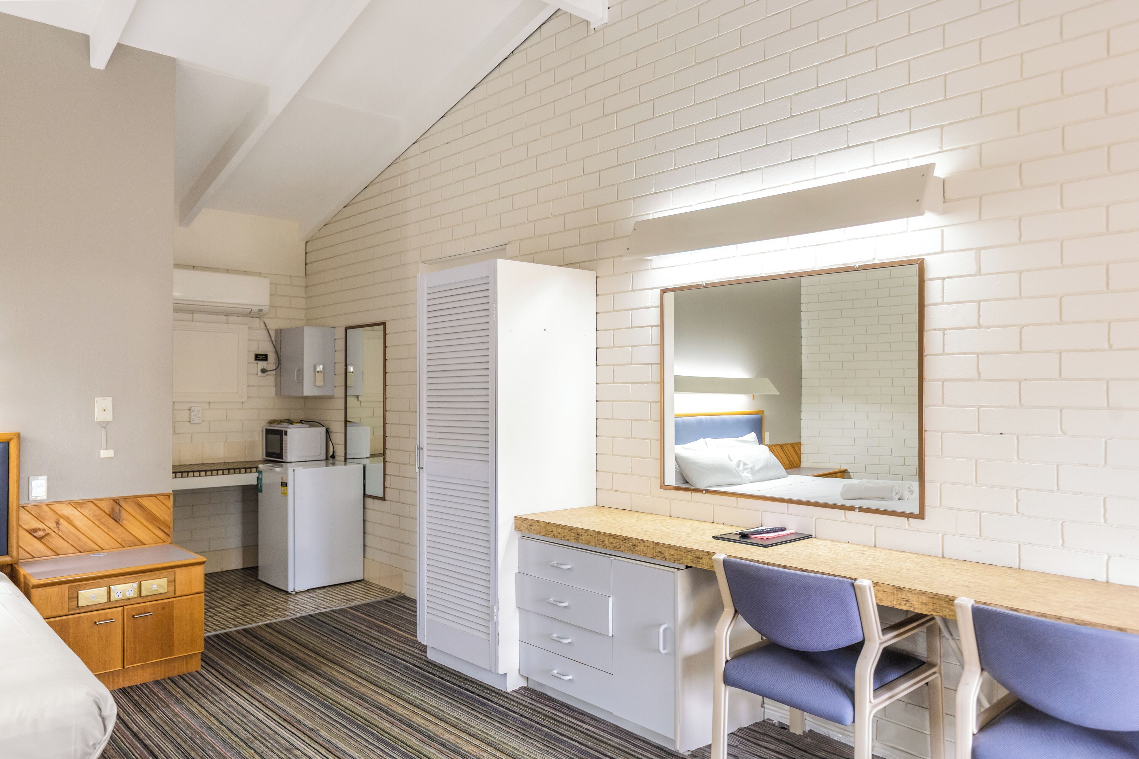 A compact hotel room with a kitchenette, a bed, a desk with two chairs, a mirror, and a striped carpet.