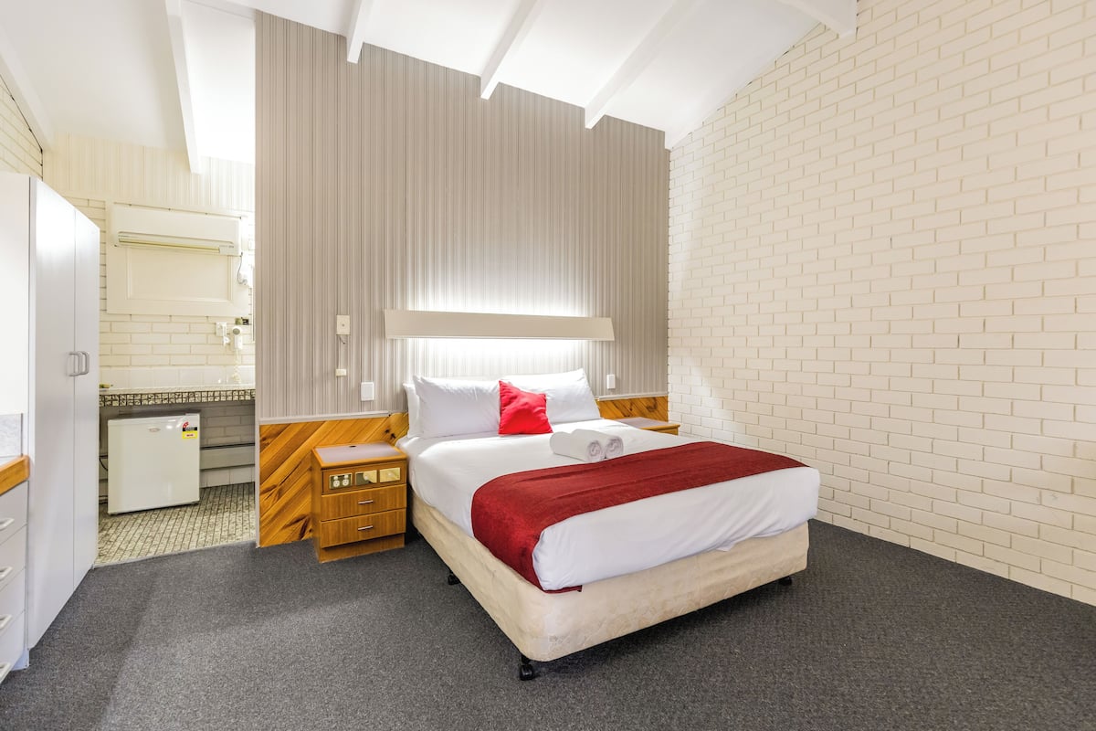 A hotel room with a bed, a wooden nightstand, a small desk, and a white brick wall.