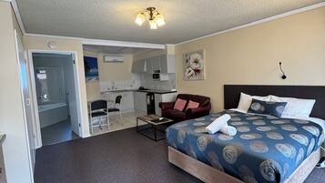 A bedroom with a large bed, a chair, a desk, and a kitchenette in the background.