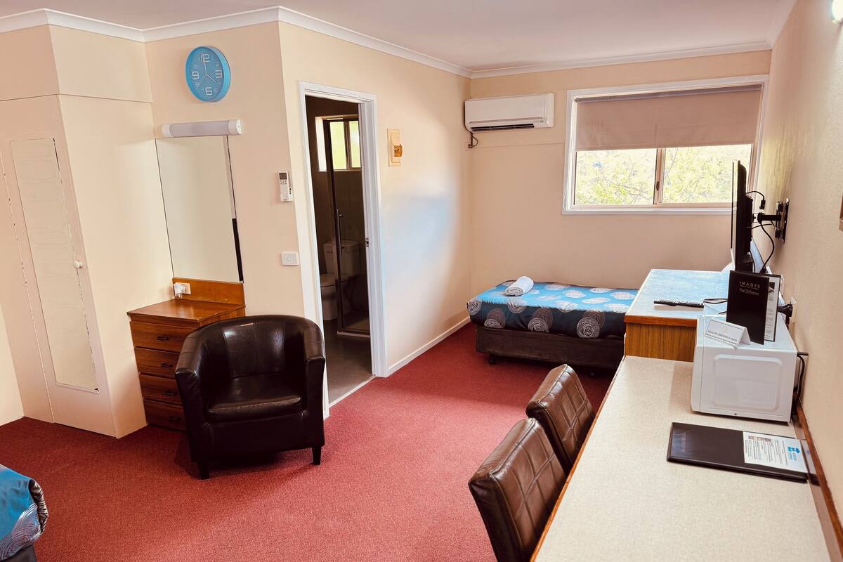 A compact room with a bed, a desk, a chair, a wardrobe, a mirror, a clock, an air conditioning unit, and a window.