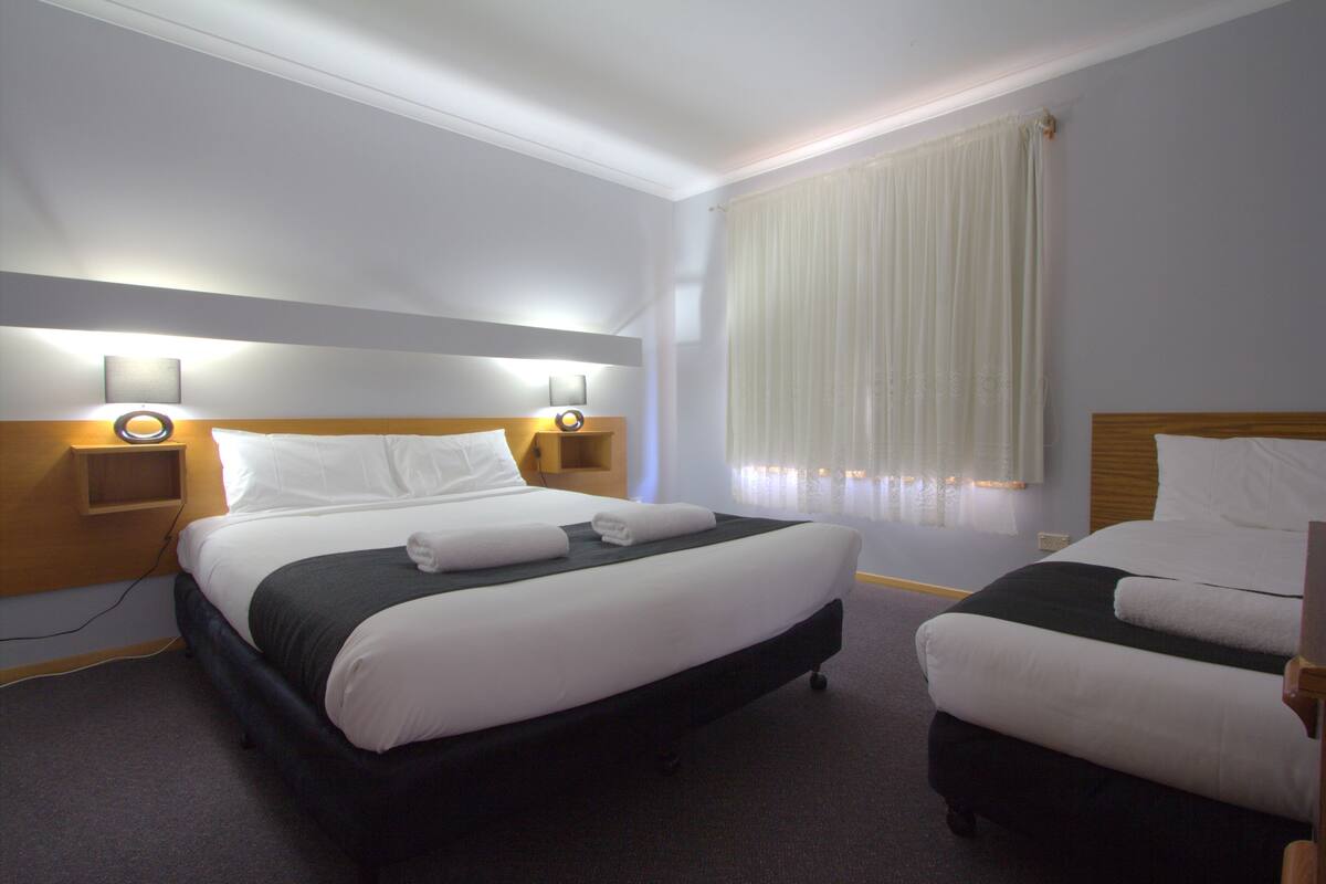 A hotel room with a double bed, a single bed, two bedside tables with lamps, a window with curtains, and a carpeted floor.