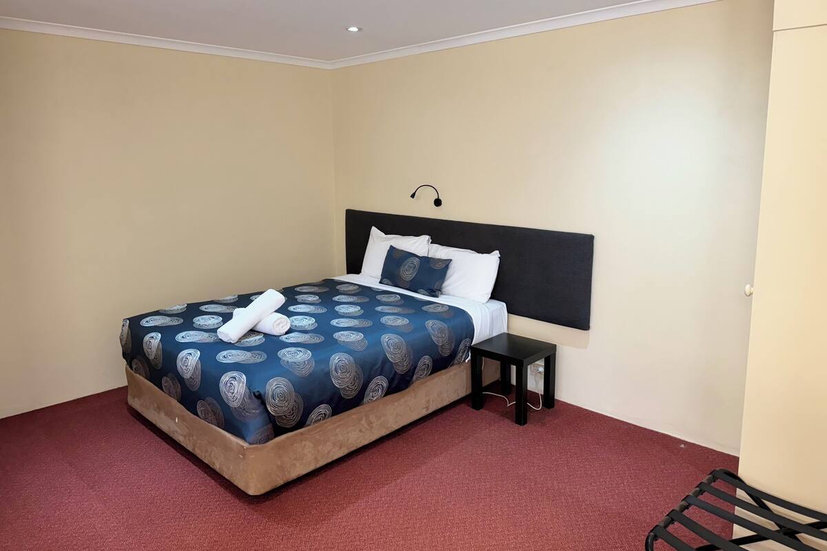 A neatly made bed with a blue patterned bedspread and white pillows, a small bedside table, and a red carpeted floor.