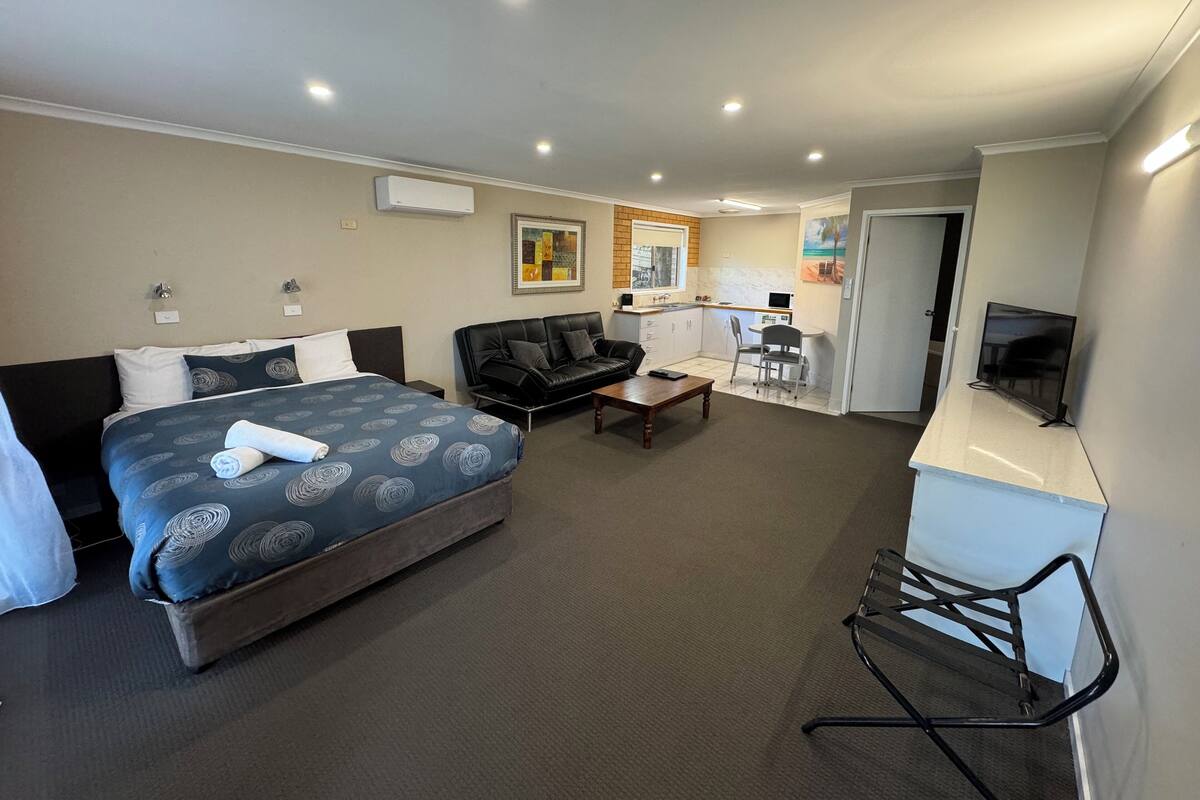 A hotel room with a large bed, a sofa, a small table, a TV, and a kitchenette.