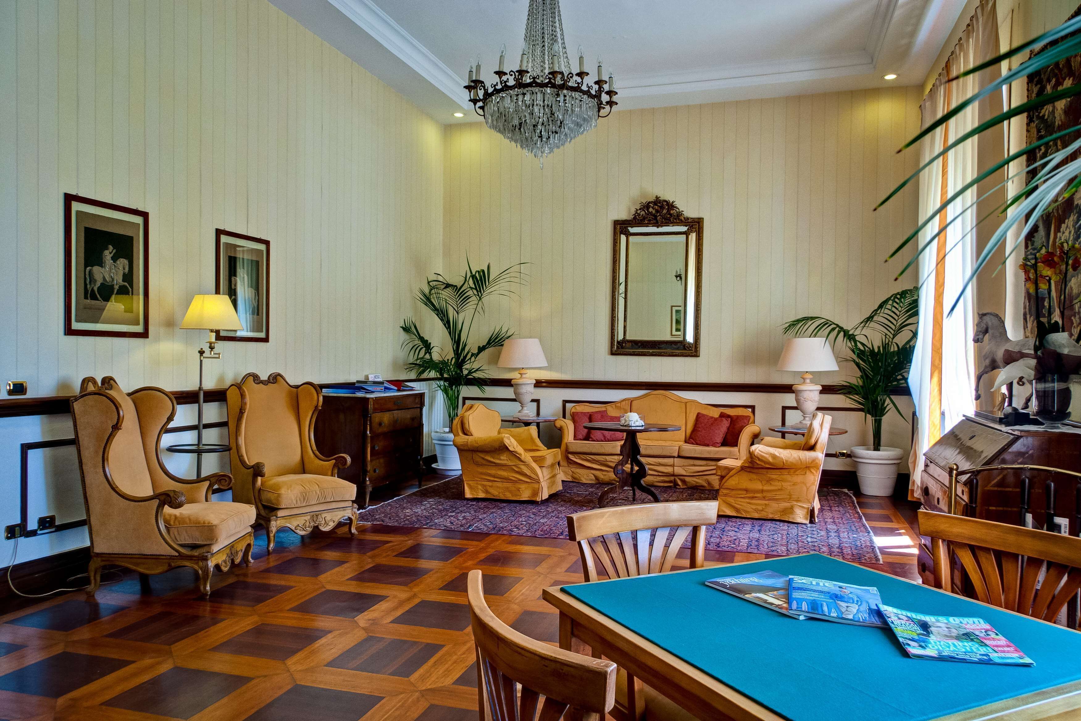 A room with a wooden parquet floor, a chandelier, a sofa set, a dining table, and a piano.