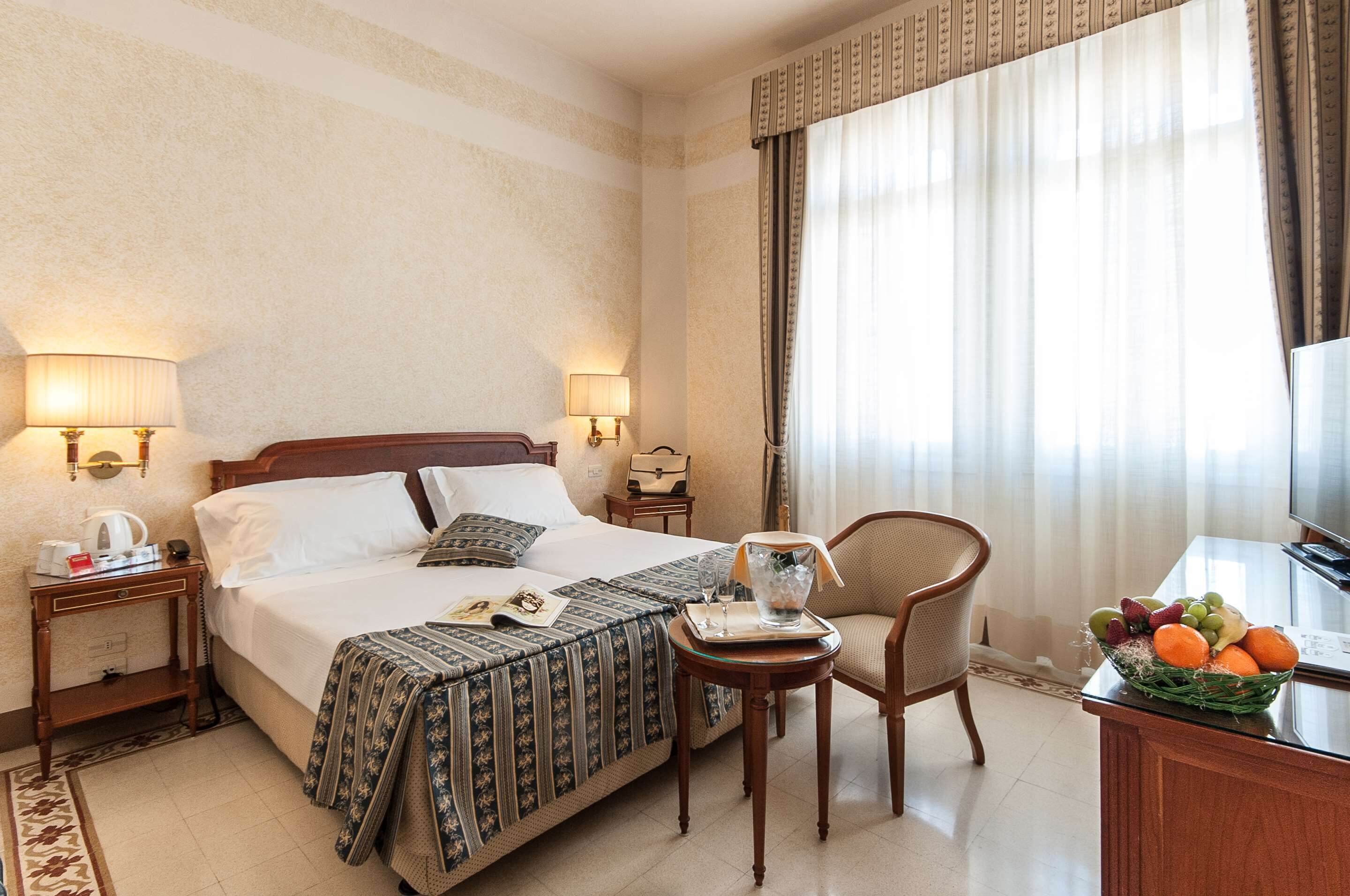Habitación de hotel con una cama grande, dos mesitas de noche con lámparas, una pequeña mesa con una cesta de fruta, una silla y un televisor.