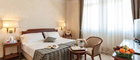 Habitación de hotel con una cama grande, dos mesitas de noche con lámparas, una pequeña mesa con una cesta de fruta, una silla y un televisor.