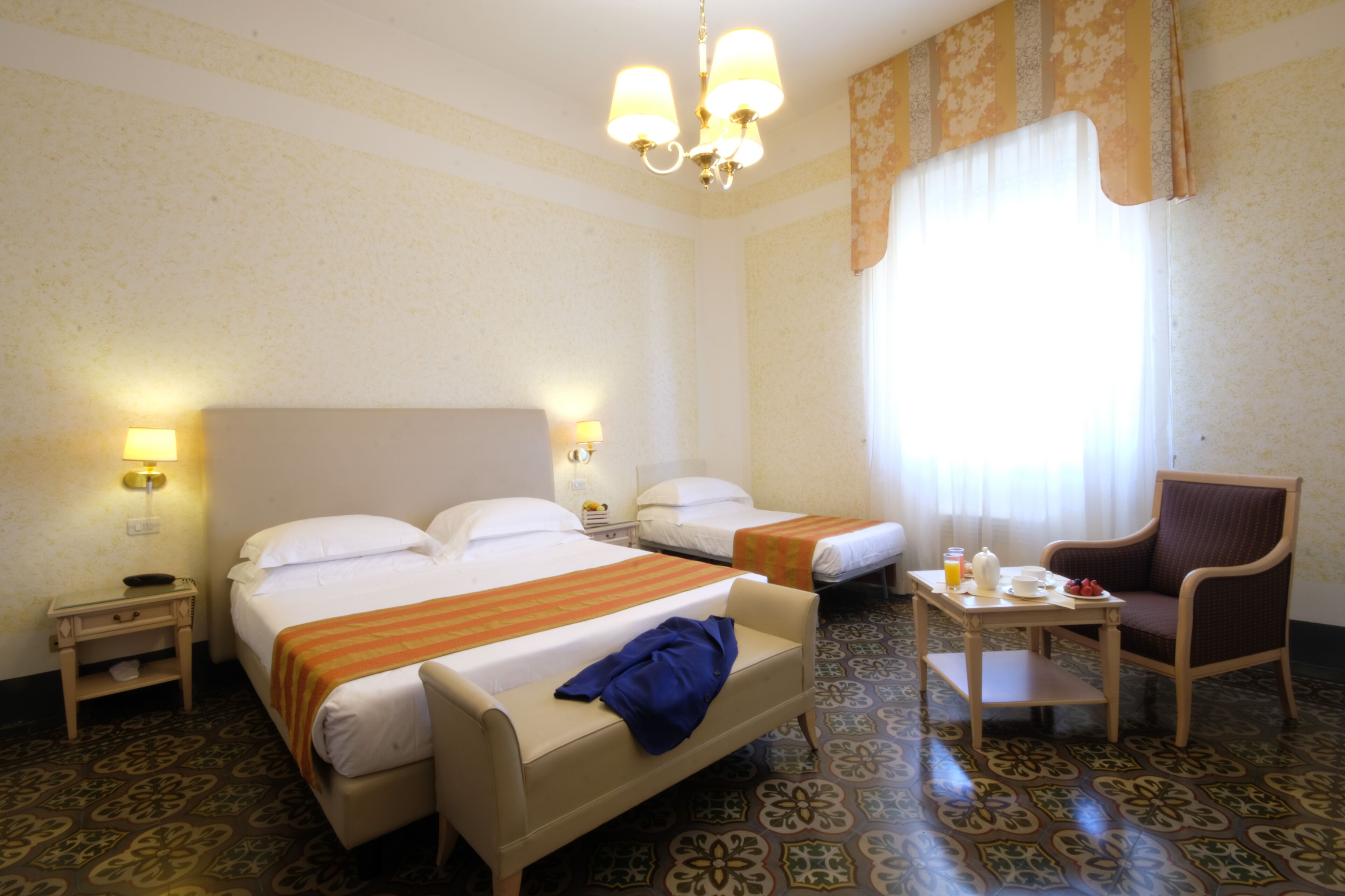 Een hotelkamer met twee bedden, een stoel, een tafeltje met een lamp en een raam met gordijnen.
