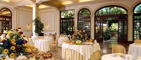 A banquet hall with round tables set for a formal event, featuring floral centerpieces and a variety of food items.
