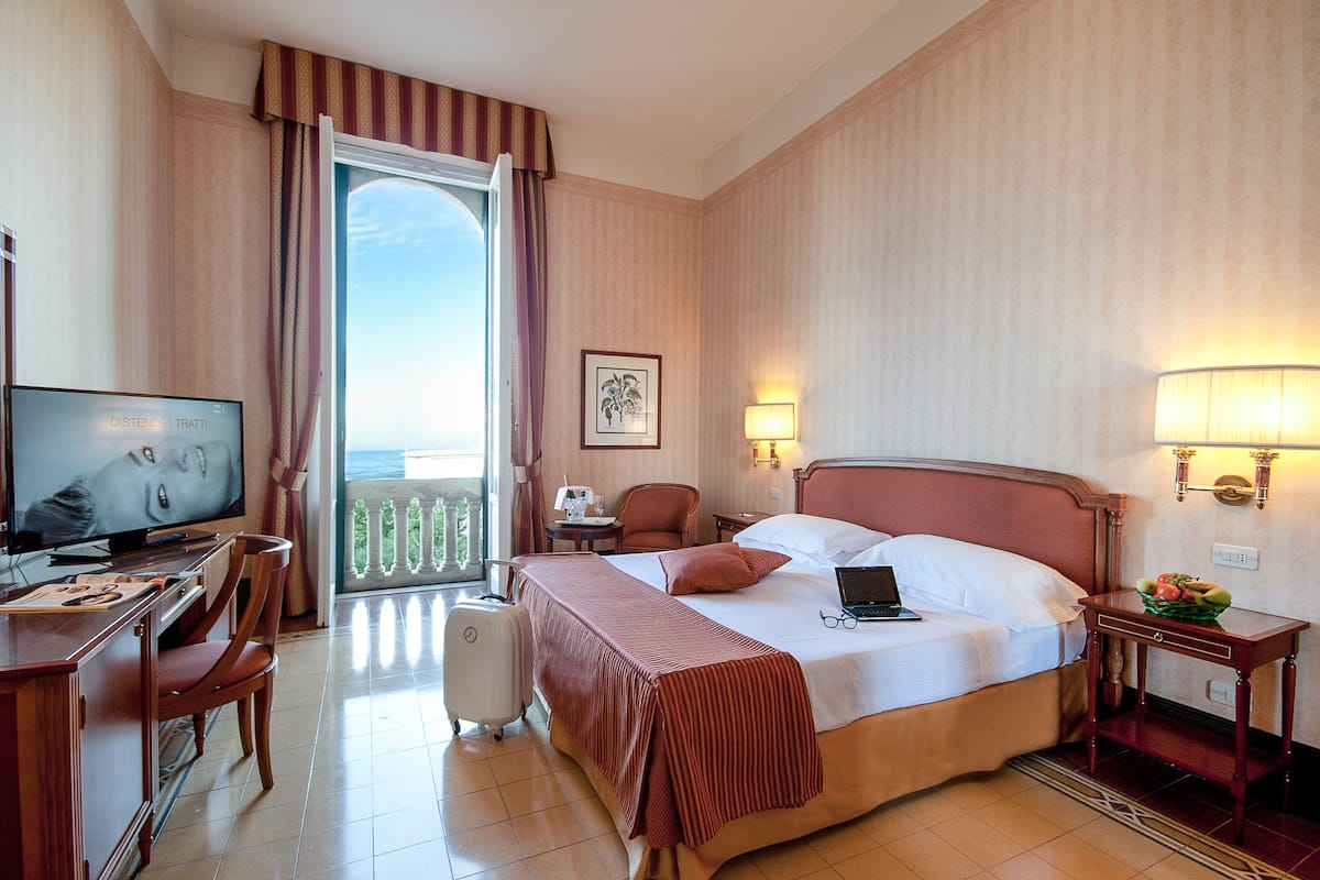 A hotel room with a bed, a desk with a computer, a television, a chair, a suitcase, and a balcony with a railing.