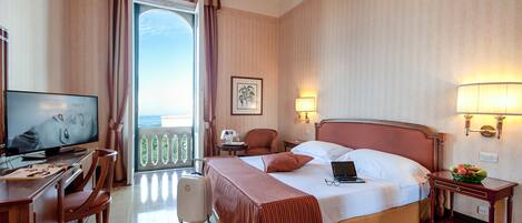 A hotel room with a bed, a desk with a computer, a television, a chair, a suitcase, and a balcony with a railing.
