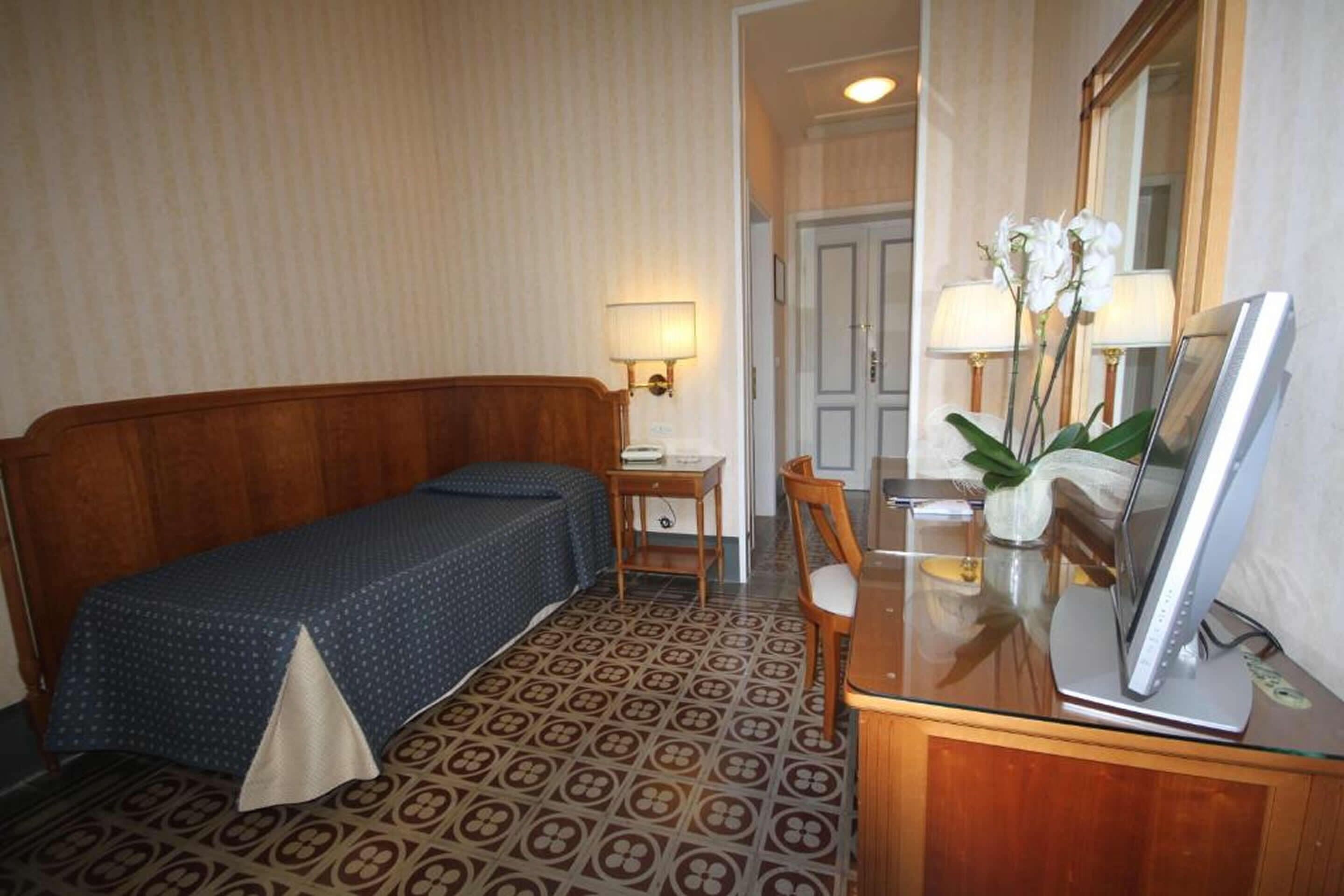 Habitación de hotel con una cama de madera, un escritorio pequeño con una lámpara, un televisor sobre una mesa de vidrio y un espejo en la pared.