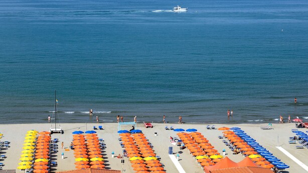 A beach with rows of umbrellas and sun loungers, a boat in the water, and people swimming and sunbathing. - Grand Hotel Royal (Viareggio)