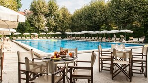 A poolside area with lounge chairs and umbrellas, a dining table with breakfast, and a swimming pool surrounded by trees. - Grand Hotel Royal (Viareggio)