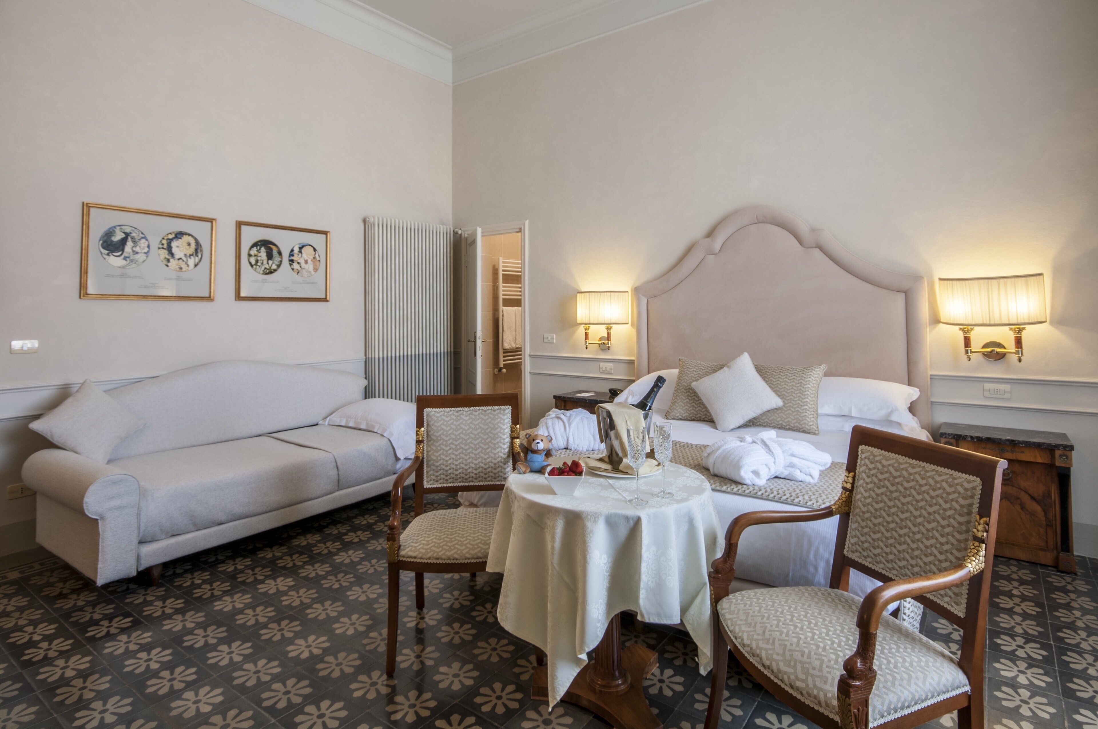 A hotel room with a bed, a sofa, two armchairs, a small round table with a white tablecloth, and two wall-mounted lamps.