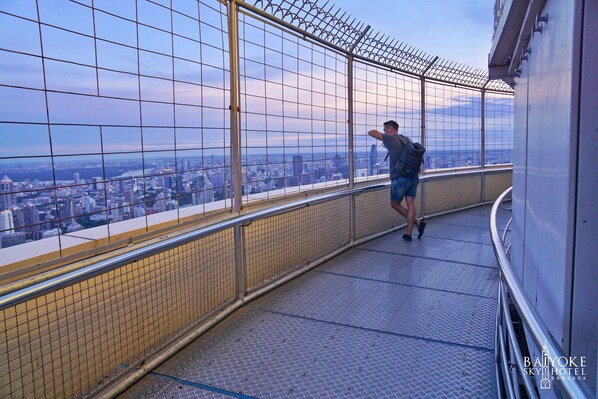 Una persona está de pie en un balcón con vista a la ciudad, barandilla de metal y un diseño de ventanas tipo celosía.