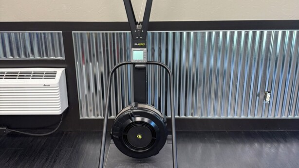 A ceiling-mounted exercise machine with a digital display, a wall-mounted air conditioner, and a window with vertical blinds.