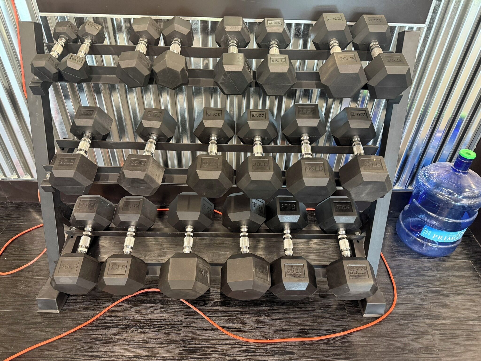 A rack of hexagonal dumbbells with weight markings, a water bottle, and orange cables on a wooden floor.