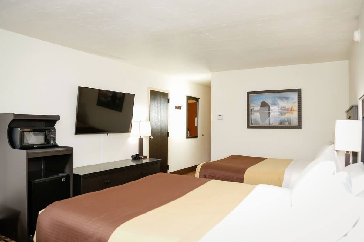 A hotel room with two beds, a TV, a desk with a microwave, a lamp, and a painting on the wall.