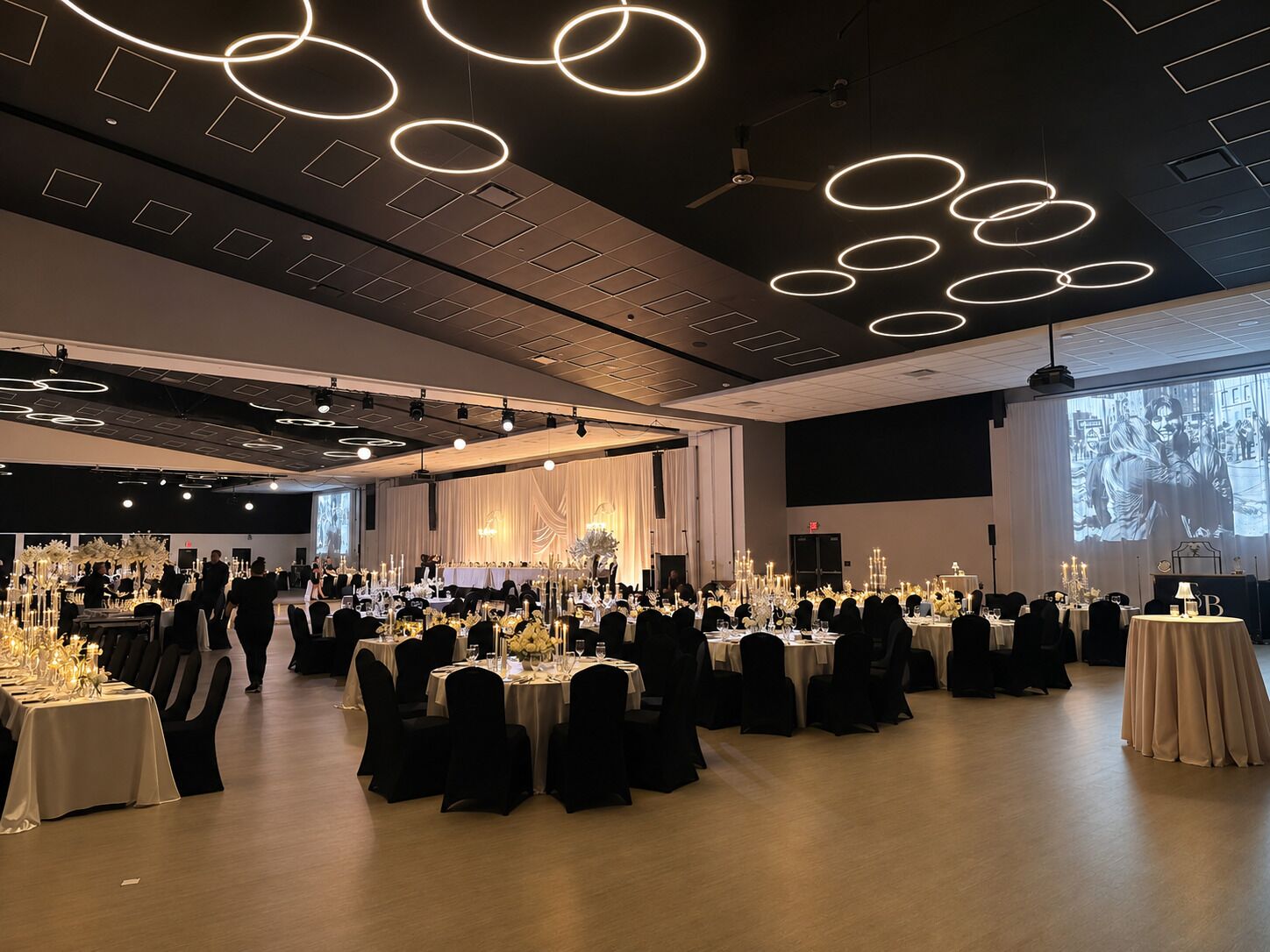 A banquet hall with round tables, chairs, and a stage for presentations.