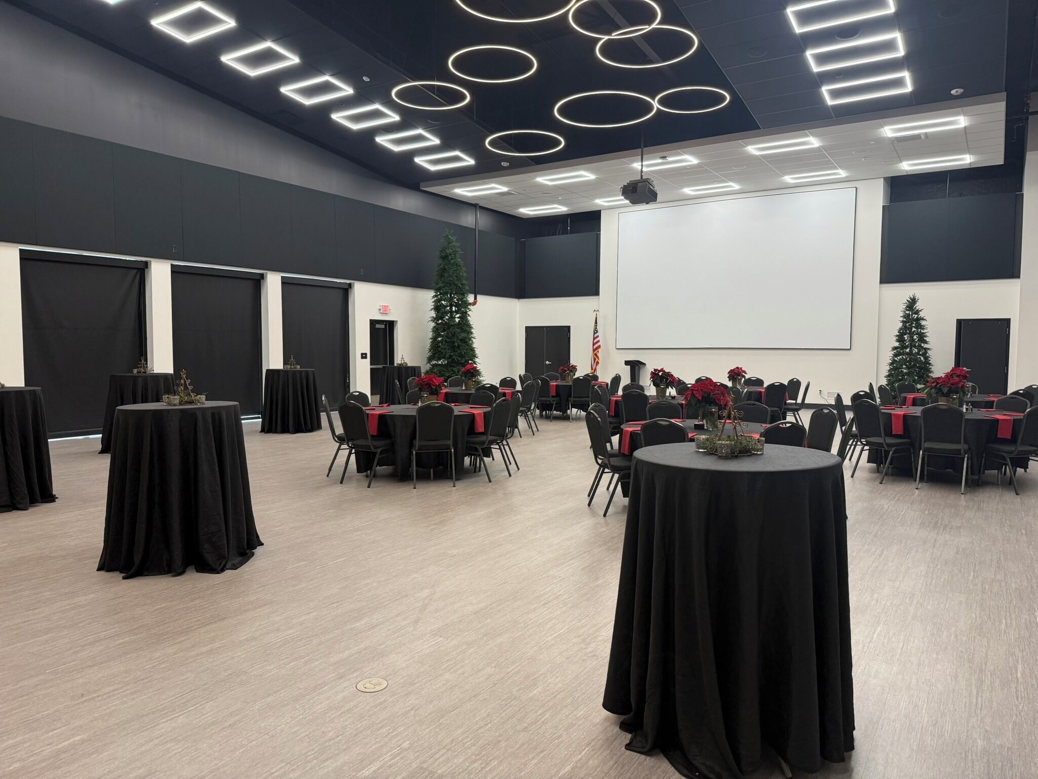Un salón de conferencias con mesas redondas, manteles negros y posavasos rojos.