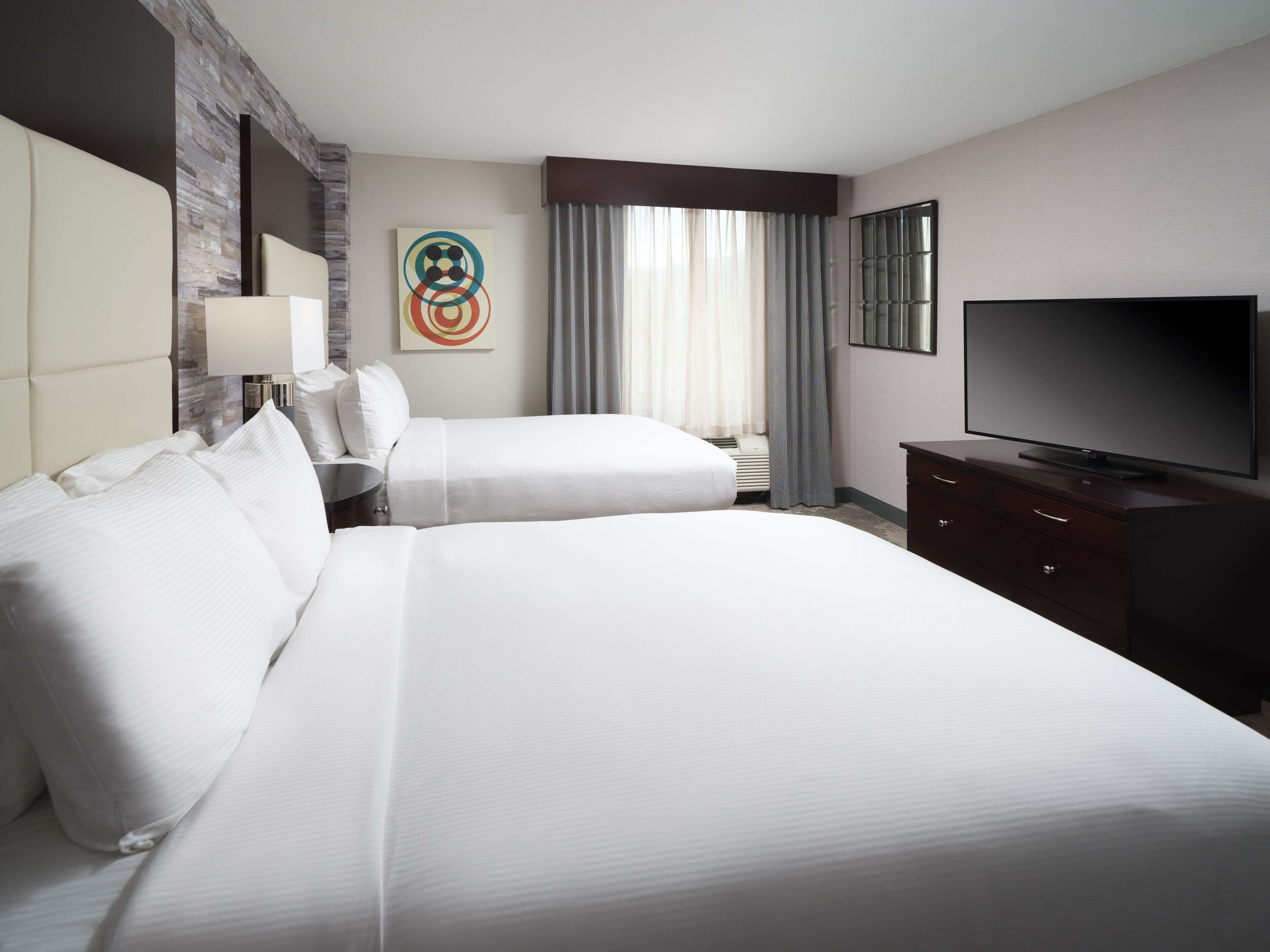 A hotel room with two beds, a flat-screen TV, a desk, and a modern art piece on the wall.