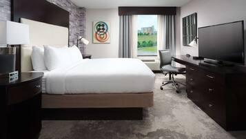 A modern hotel room with a large bed, a desk, a flat-screen TV, and a view of a green landscape through the window.