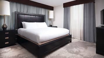 A modern hotel room with a large bed, dark wood headboard, two bedside tables with lamps, and a large window with curtains.