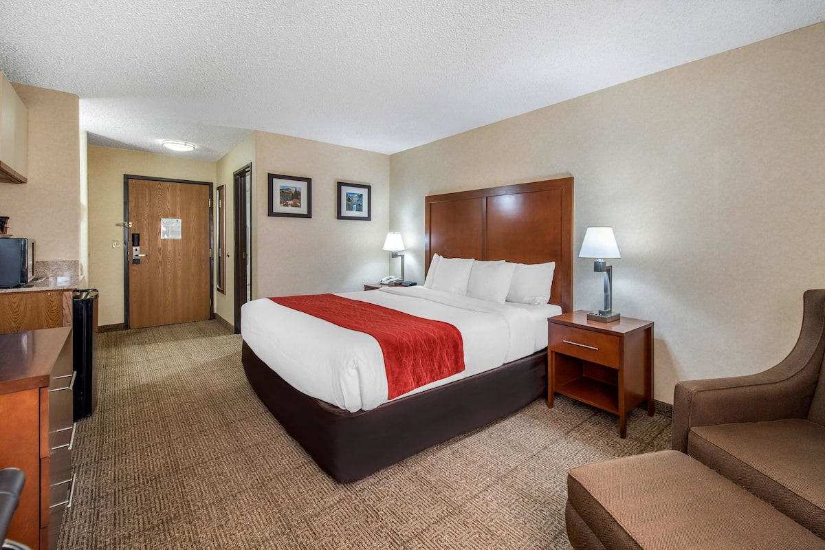 A hotel room with a large bed, two bedside tables with lamps, a wooden headboard, a brown armchair, and a desk with a computer.