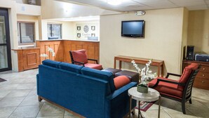 Un salon avec un canapé bleu, des fauteuils rouges et noirs, une petite table sur laquelle est posé un vase de fleurs, et un téléviseur fixé au mur.