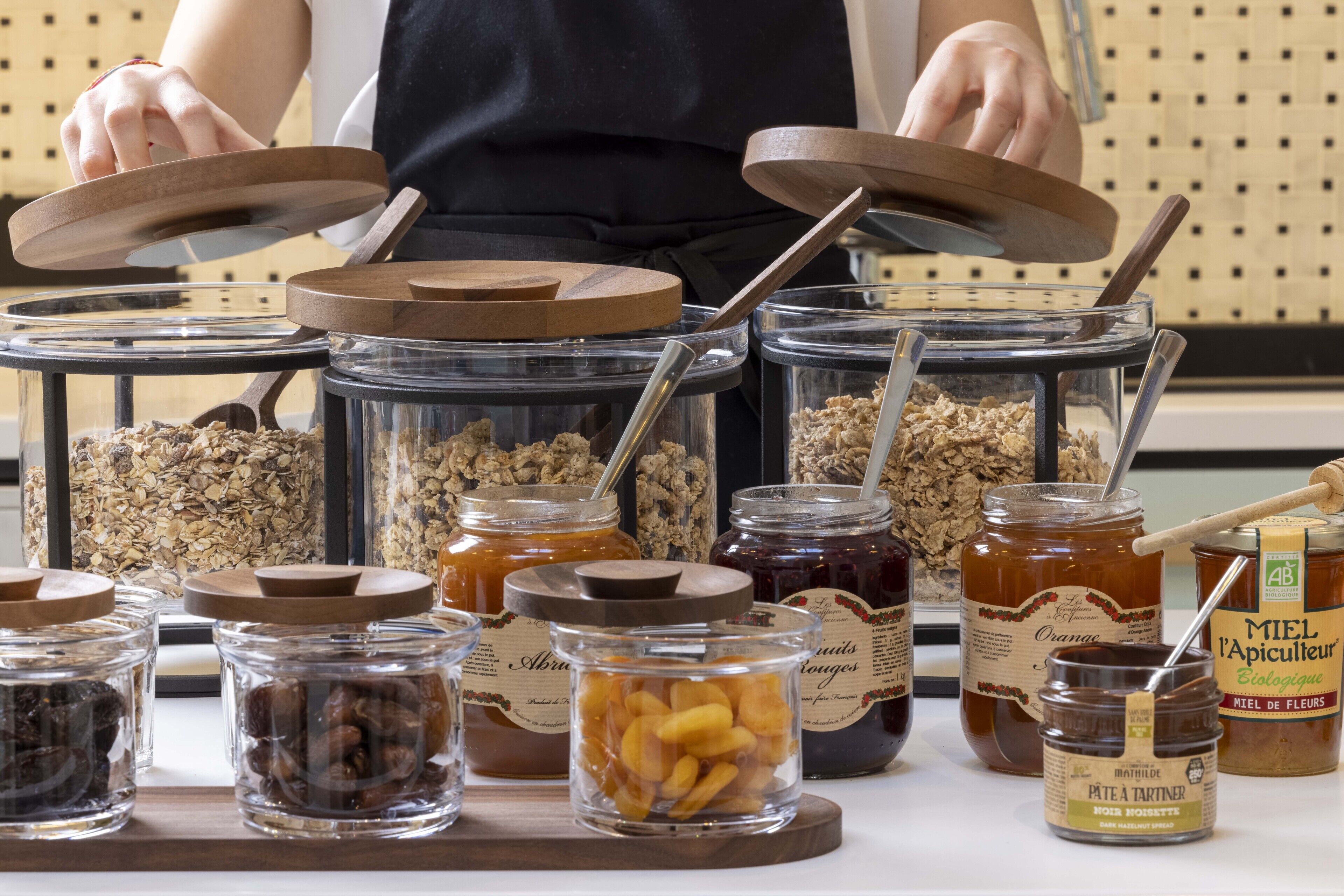Verschillende soorten levensmiddelen in glazen potten met houten deksels, zoals granola, gedroogd fruit en honing.