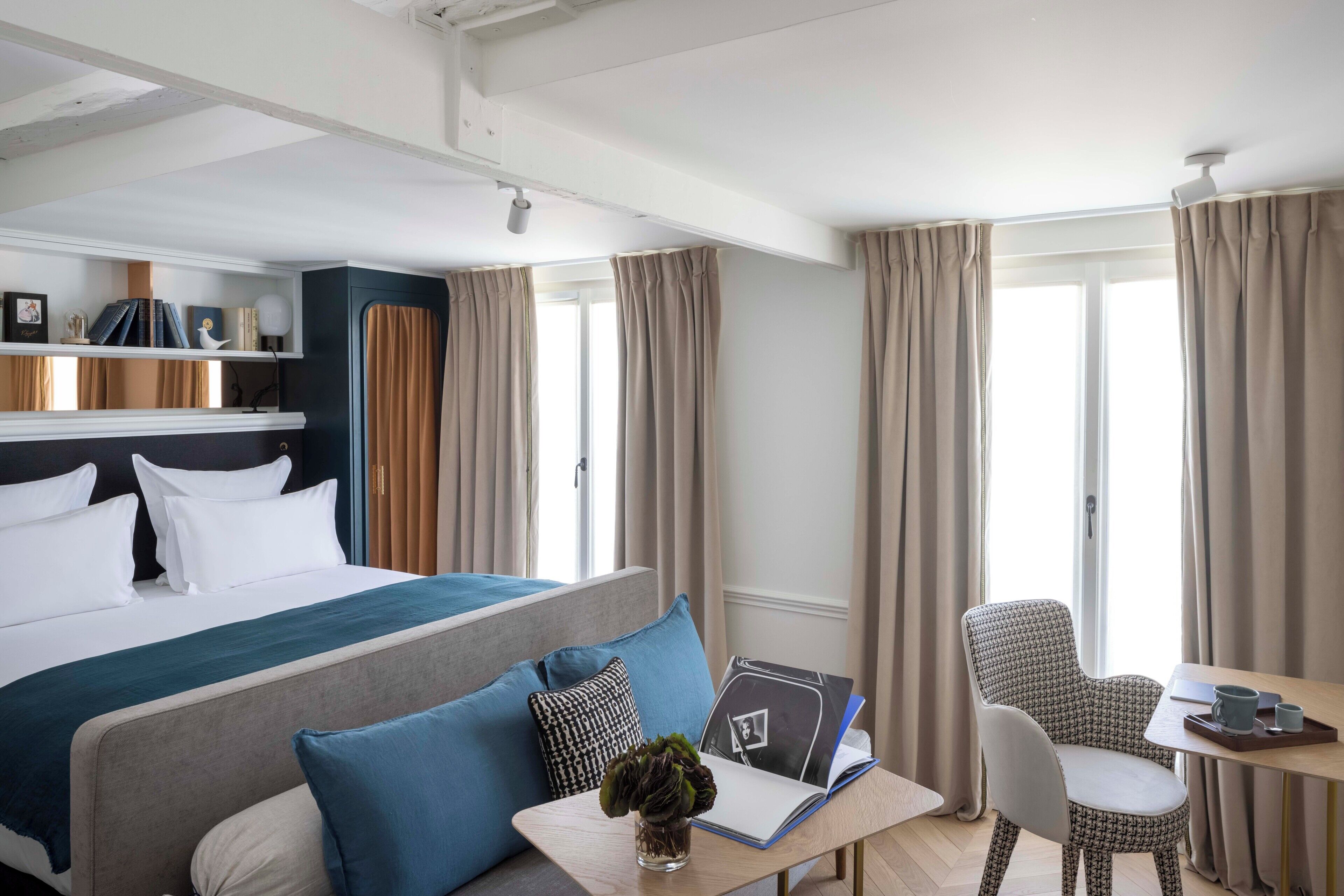A hotel room with a bed, a desk with a chair, a bookshelf, and a window with curtains.