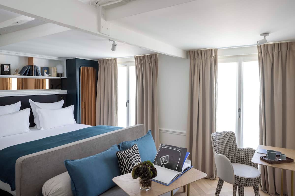 A hotel room with a bed, a desk with a chair, a bookshelf, and a window with curtains.