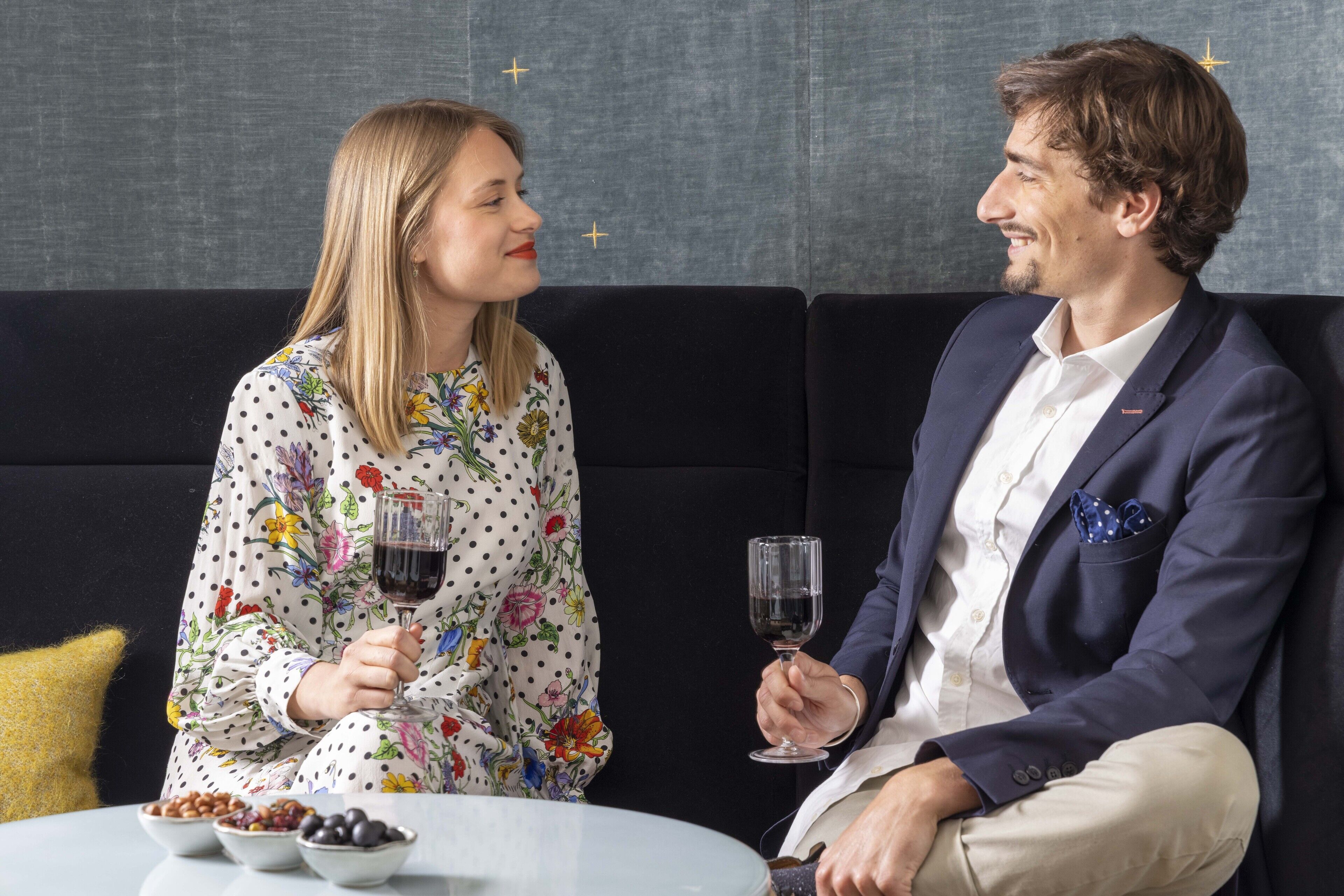 Deux personnes sont assises sur un canapé noir, une table les séparant, et elles tiennent chacune un verre de vin.