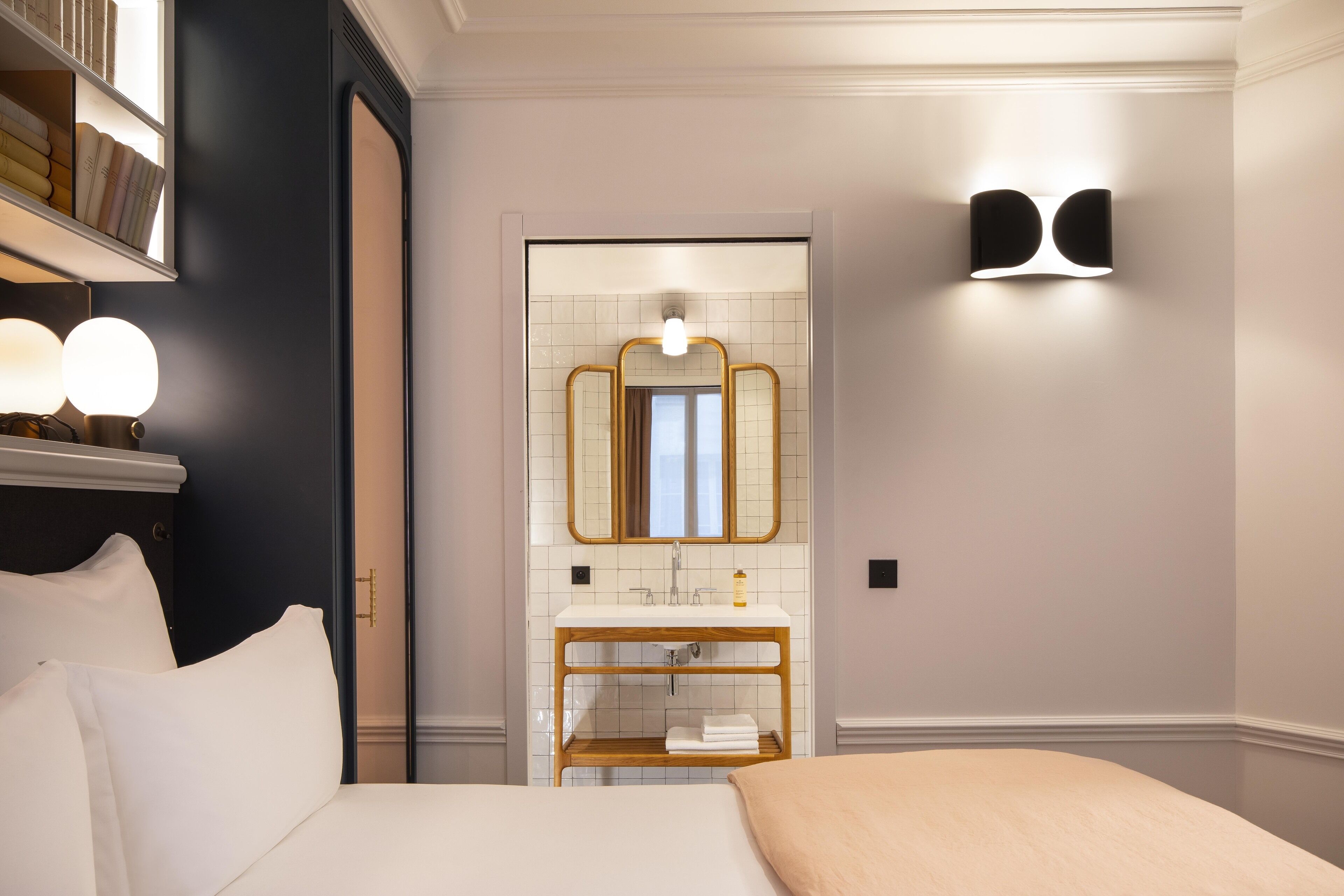 A hotel room with a bed, bedside lamps, a mirror, a vanity with a sink, and a shelf with towels.
