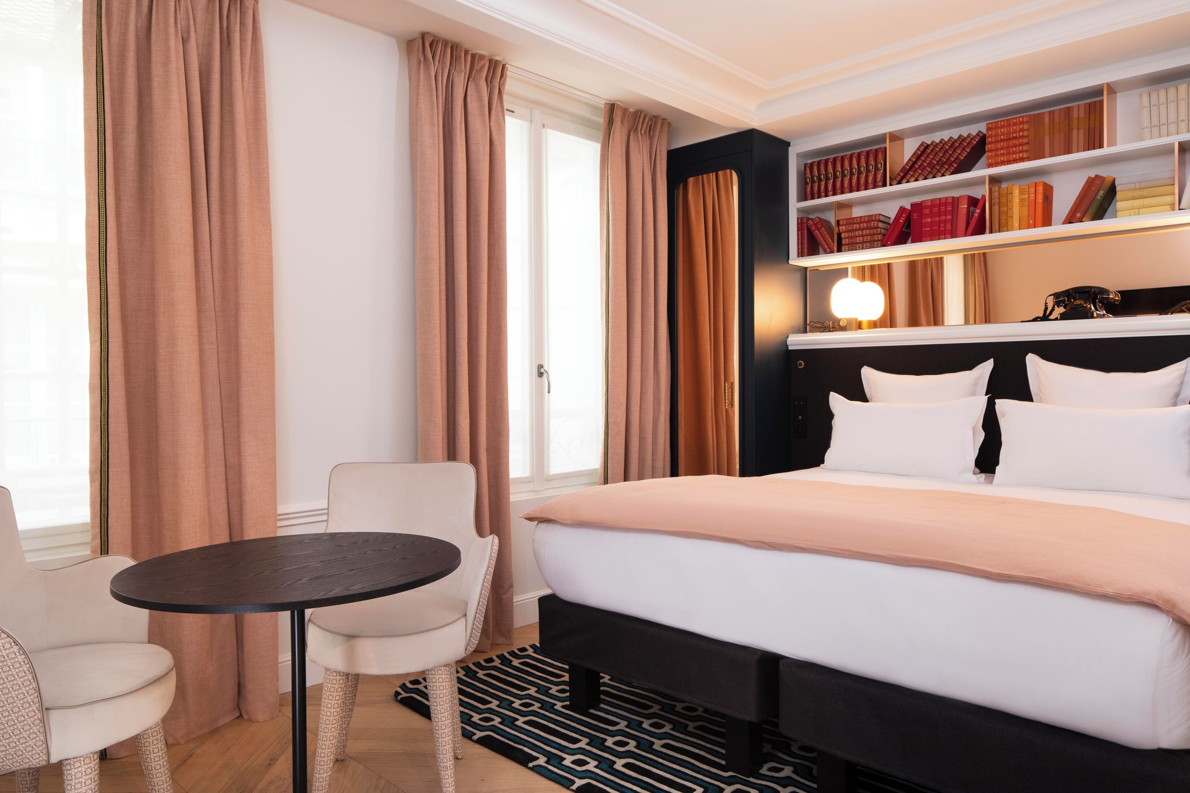 A hotel room with a bed, a bedside table, a chair, a bookshelf, and a window with curtains.