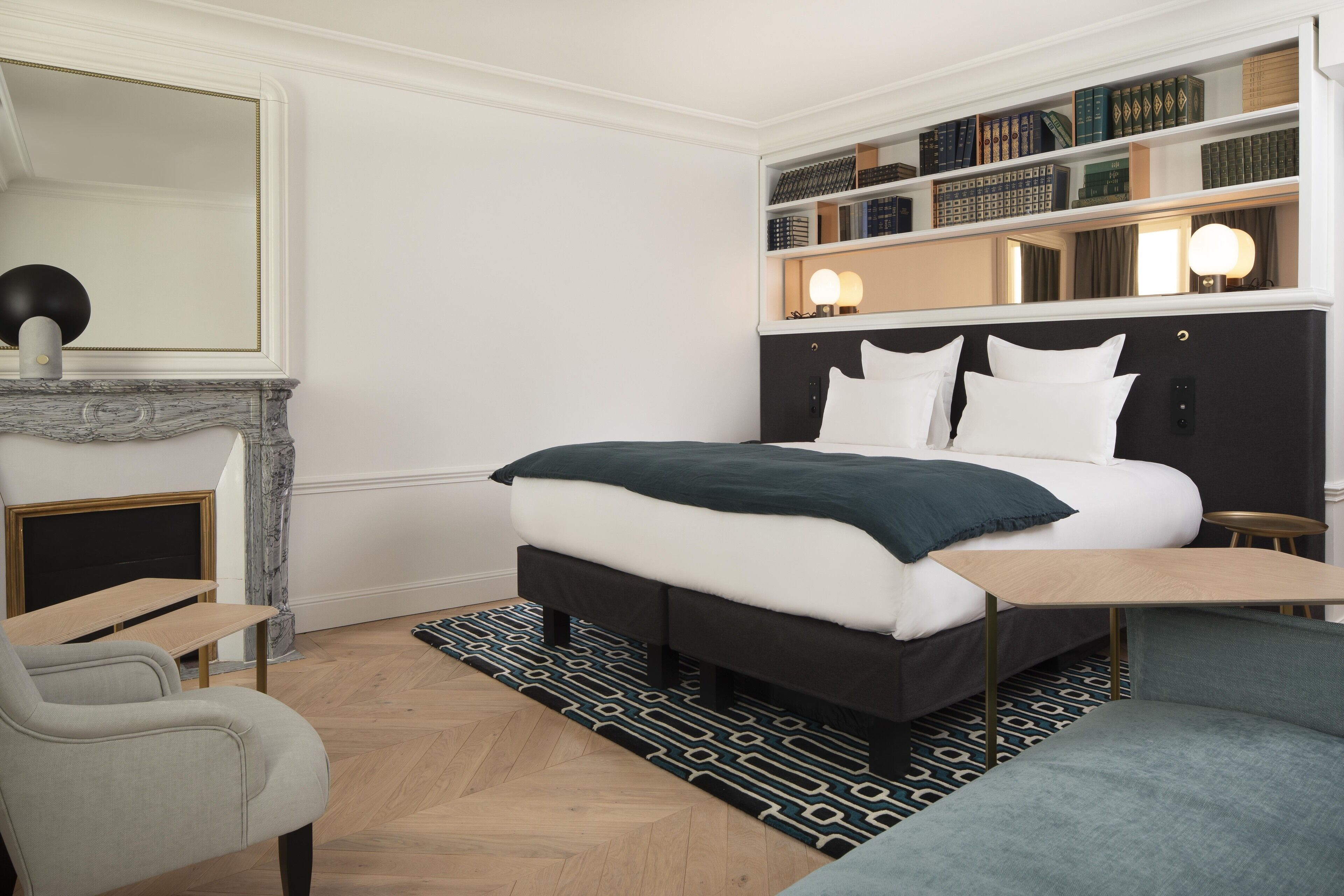 A modern bedroom with a large bed, a bedside table, a chair, a bookshelf, and a fireplace.