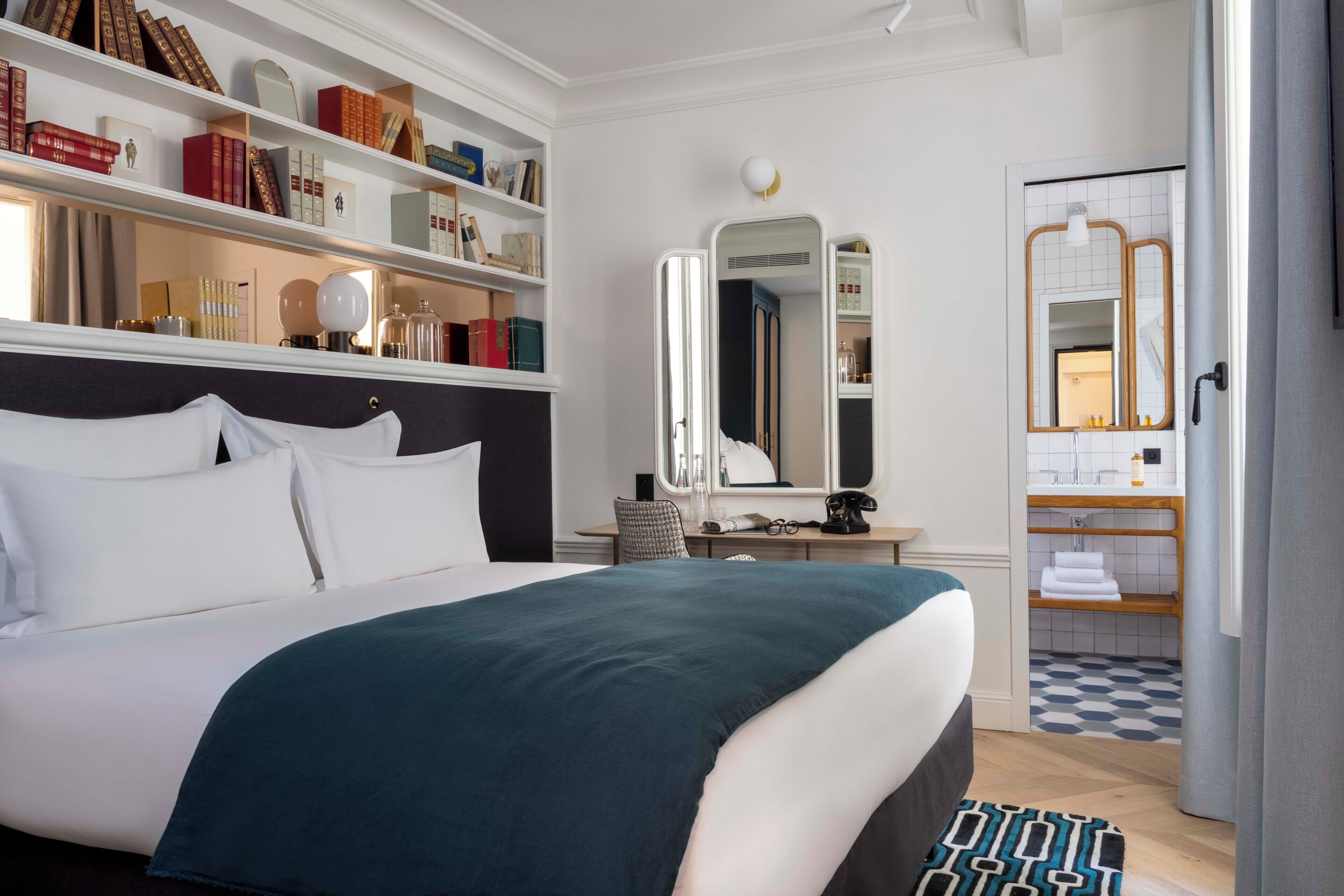 A hotel room with a large bed, a bookshelf, a desk with a chair, a mirror, and a bathroom visible through an open door.