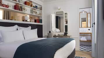 A hotel room with a large bed, a desk with a chair, a bookshelf, a bathroom with a mirror, and a patterned rug.