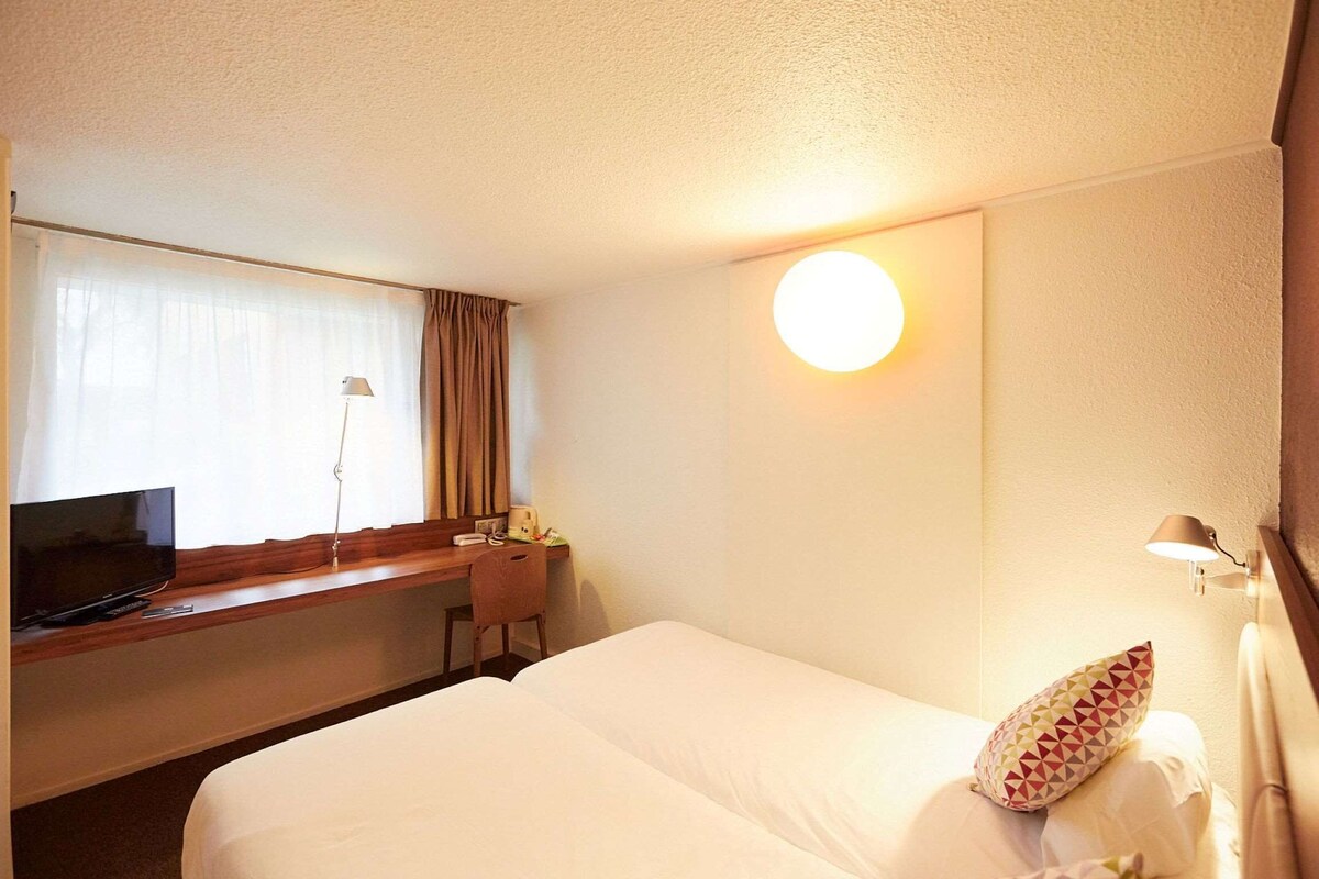 Une chambre d’hôtel avec un lit, un bureau avec une télévision, une lampe et une fenêtre avec des rideaux.