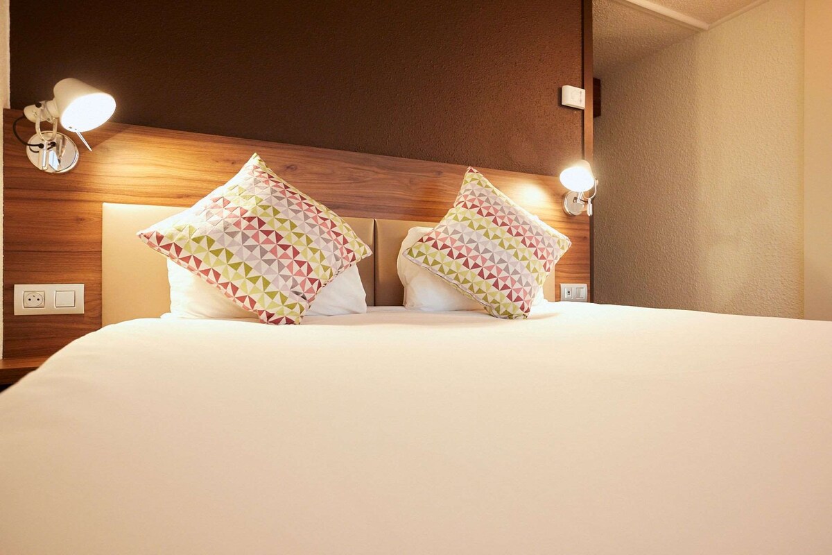Une chambre d’hôtel avec une tête de lit en bois, deux lampes de chevet et des coussins à motifs.