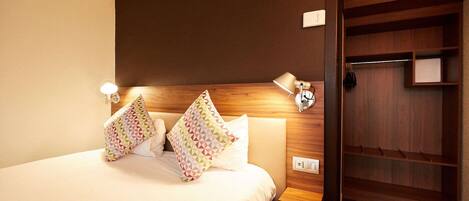 A hotel room with a bed, two patterned pillows, a nightstand with a lamp, and a wooden wardrobe with shelves.