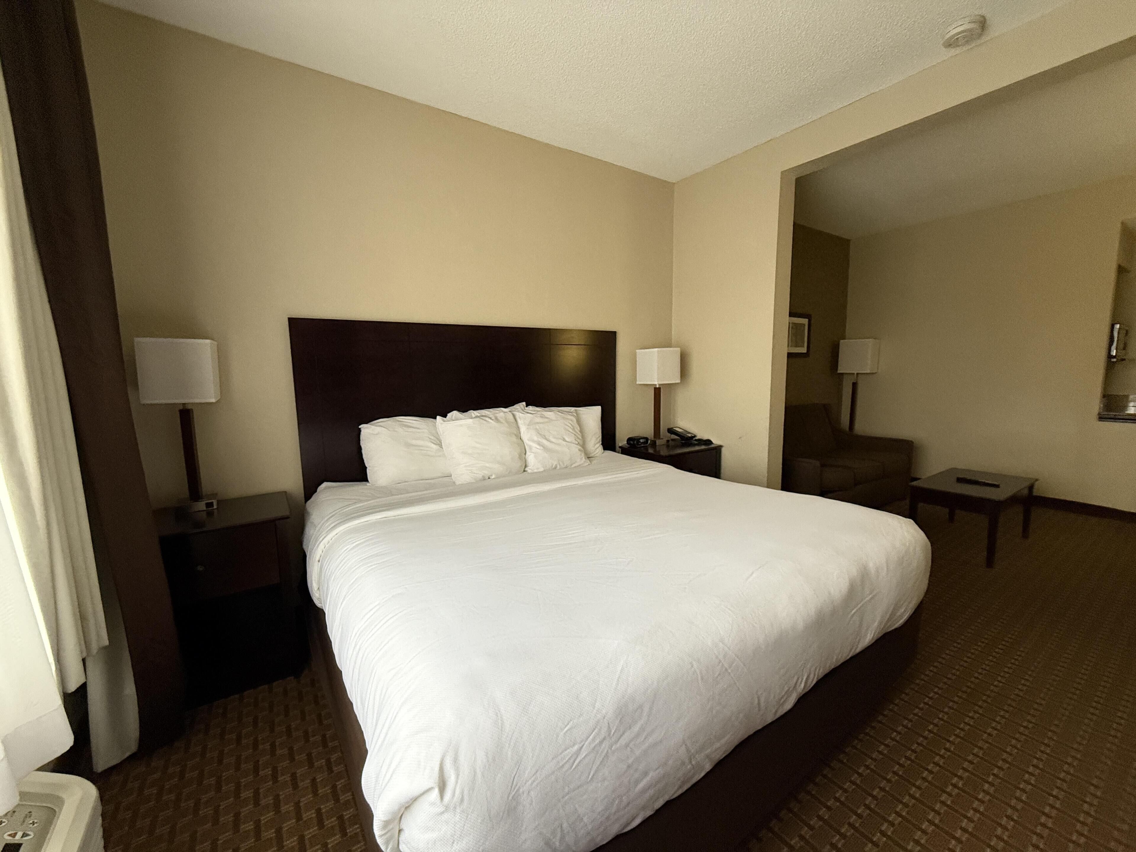 Quarto de hotel com uma cama grande, duas mesas de cabeceira com luminárias, uma cadeira e uma pequena mesa.