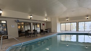 En indendørs swimmingpool med store glasvægge, en badestige og et siddeområde med borde og stole.