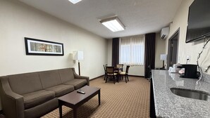 Et hotelværelse med sofa, et lille bord, et spisebord med stole, et tekøkken med vask og et fladskærms-tv monteret på væggen.