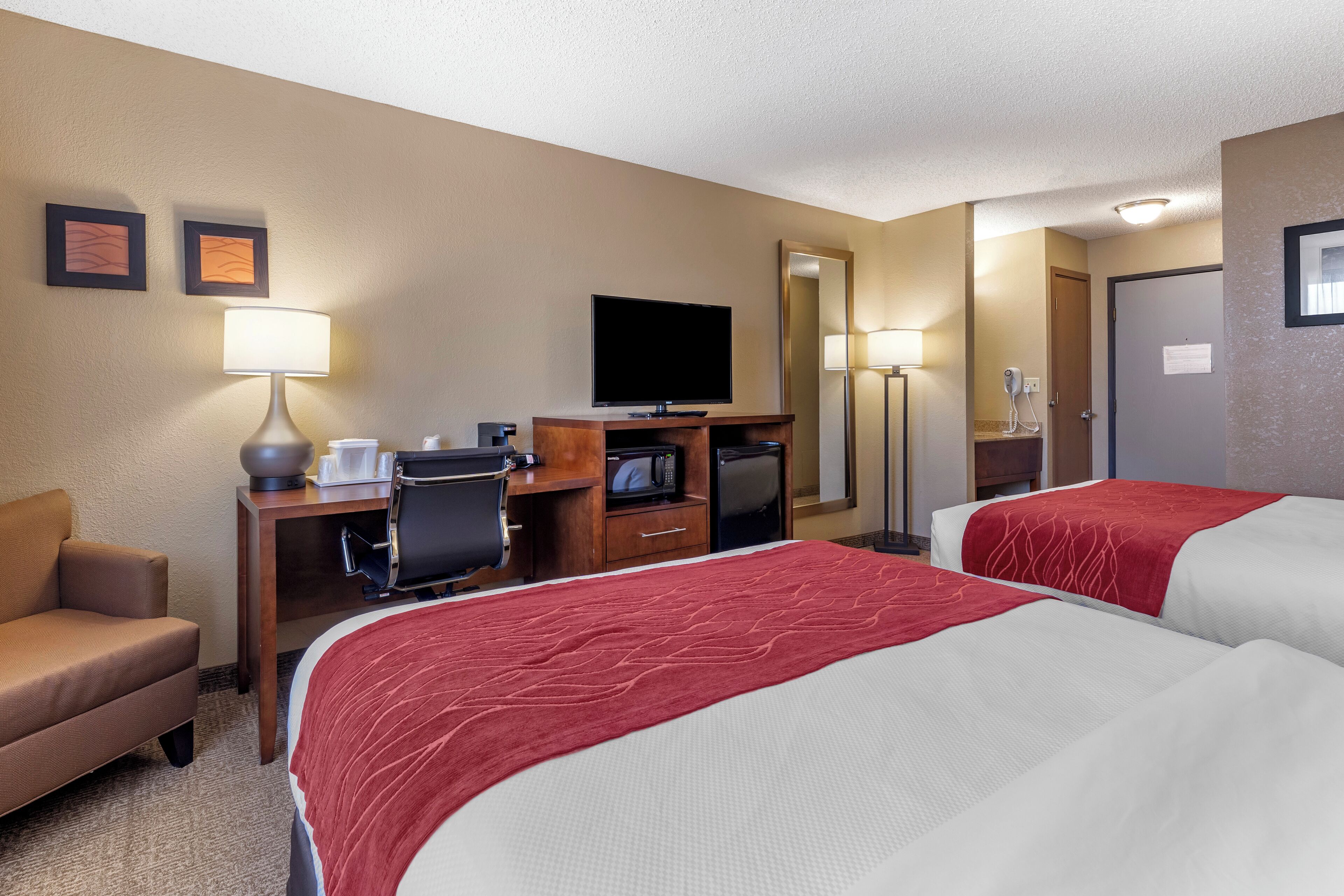 A hotel room with two beds, a desk with a TV, a lamp, a chair, and a microwave.