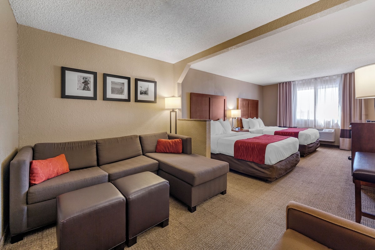 A hotel room with a sectional sofa, a bed with a red blanket, a desk, and two framed pictures on the wall.